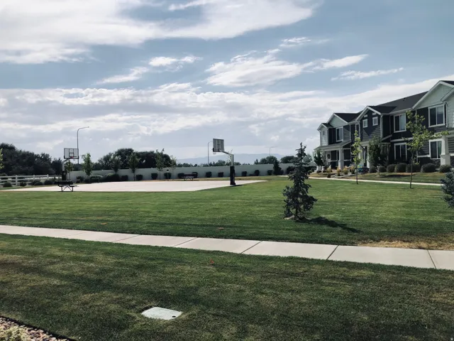 Community green space featuring a concrete basketball court with two hoops, surrounded by a white fence and mature trees