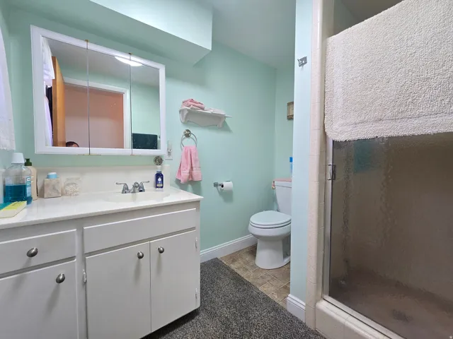 Bathroom featuring a white vanity with integrated sink, white cabinetry with silver hardware, a mirrored medicine cabinet, a toilet, and a glass-enclosed shower