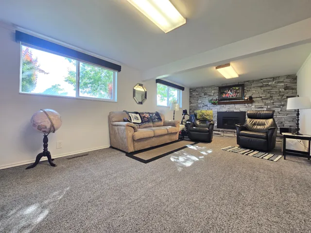 Spacious living area featuring plush gray carpeting, multiple windows, a stone-faced fireplace with a dark mantel, and recessed ceiling lighting