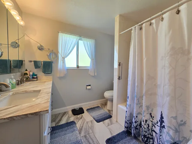 Bathroom featuring a vanity with an integrated sink, a light-toned countertop, a window with privacy curtains, a toilet, and a shower/tub combination with a grab bar
