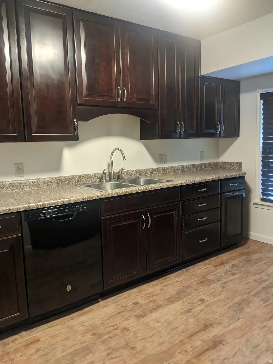 Kitchen featuring dark wood-finish cabinetry with silver-tone hardware, a double basin stainless steel sink, speckled countertops, and wood-finish flooring