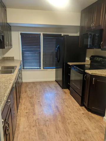 Galley kitchen featuring wood-finish flooring, dark wood cabinetry, granite-style countertops, and a full suite of black appliances