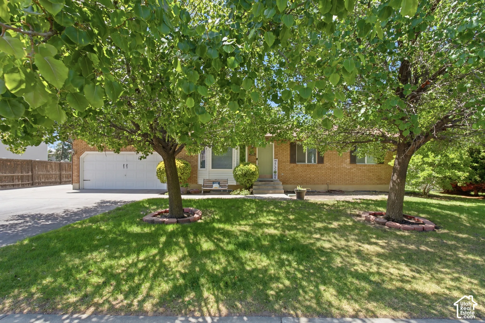 Brick facade residence featuring a two-car garage, mature front yard trees, a paved driveway, and a fenced side yard