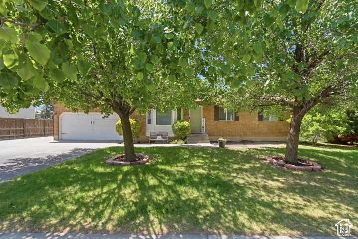 Brick facade residence featuring a two-car garage, mature front yard trees, a paved driveway, and a fenced side yard