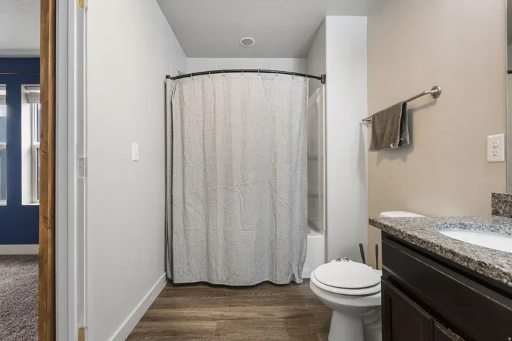 Full bathroom featuring wood-finish flooring, a white tub and shower combination with a curved shower rod, a white toilet, and a vanity with a speckled countertop and dark wood-finish cabinetry