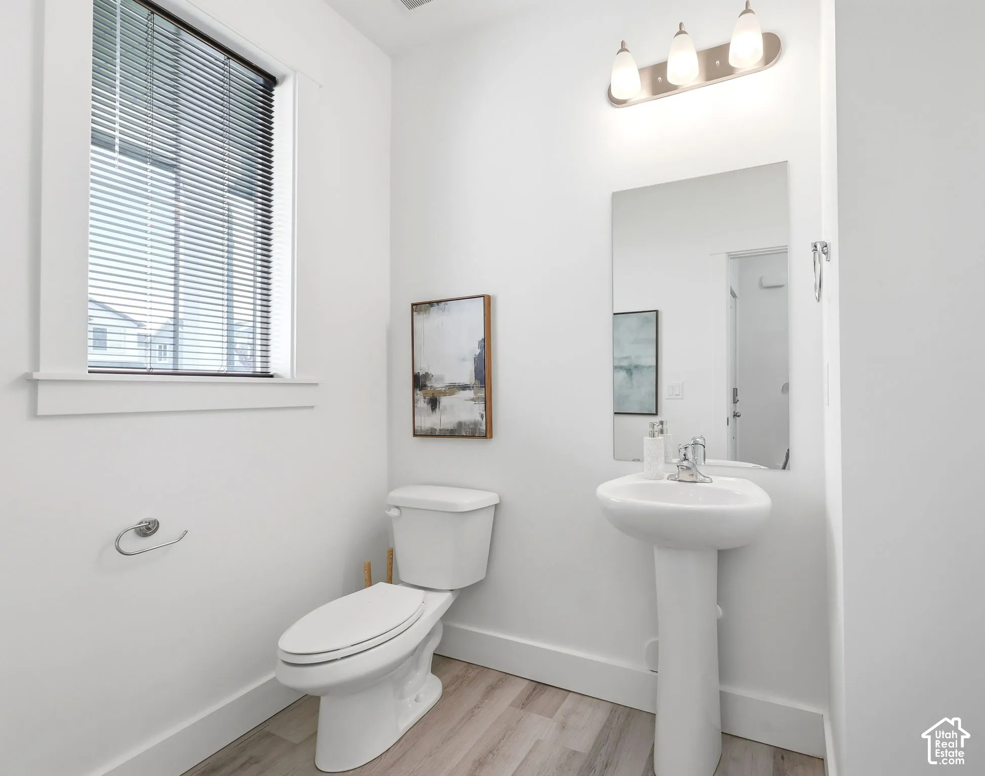Bathroom featuring a pedestal sink, wall-mounted mirror, three-light vanity fixture, and wood-finish flooring
