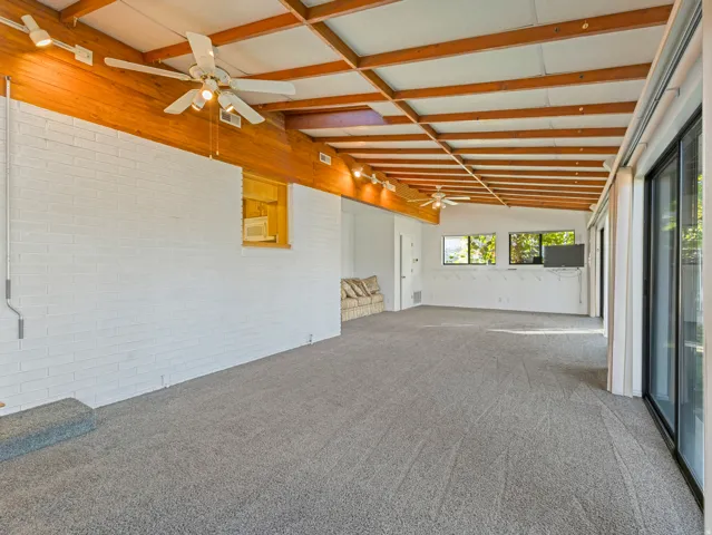 Spacious room with exposed wood beam ceiling, white painted brick wall, gray carpet flooring, and multiple ceiling fans