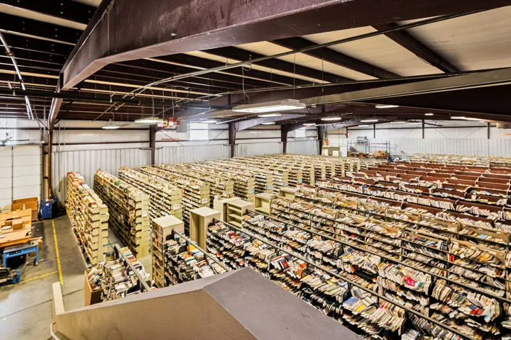 Spacious warehouse interior featuring high ceilings, exposed structural beams, extensive shelving systems, concrete flooring, and fluorescent lighting fixtures