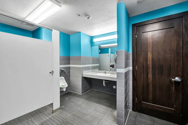 Commercial restroom featuring a dark wood-finish entry door, light blue and gray tiled walls, a vanity with an integrated sink, a rectangular mirror, and gray tile flooring