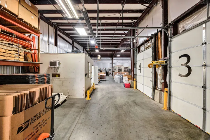 Industrial warehouse interior featuring exposed steel ceiling trusses, fluorescent lighting, and concrete flooring