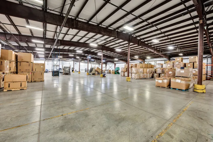 Expansive industrial interior featuring concrete flooring, exposed structural beams, high ceilings, and fluorescent lighting