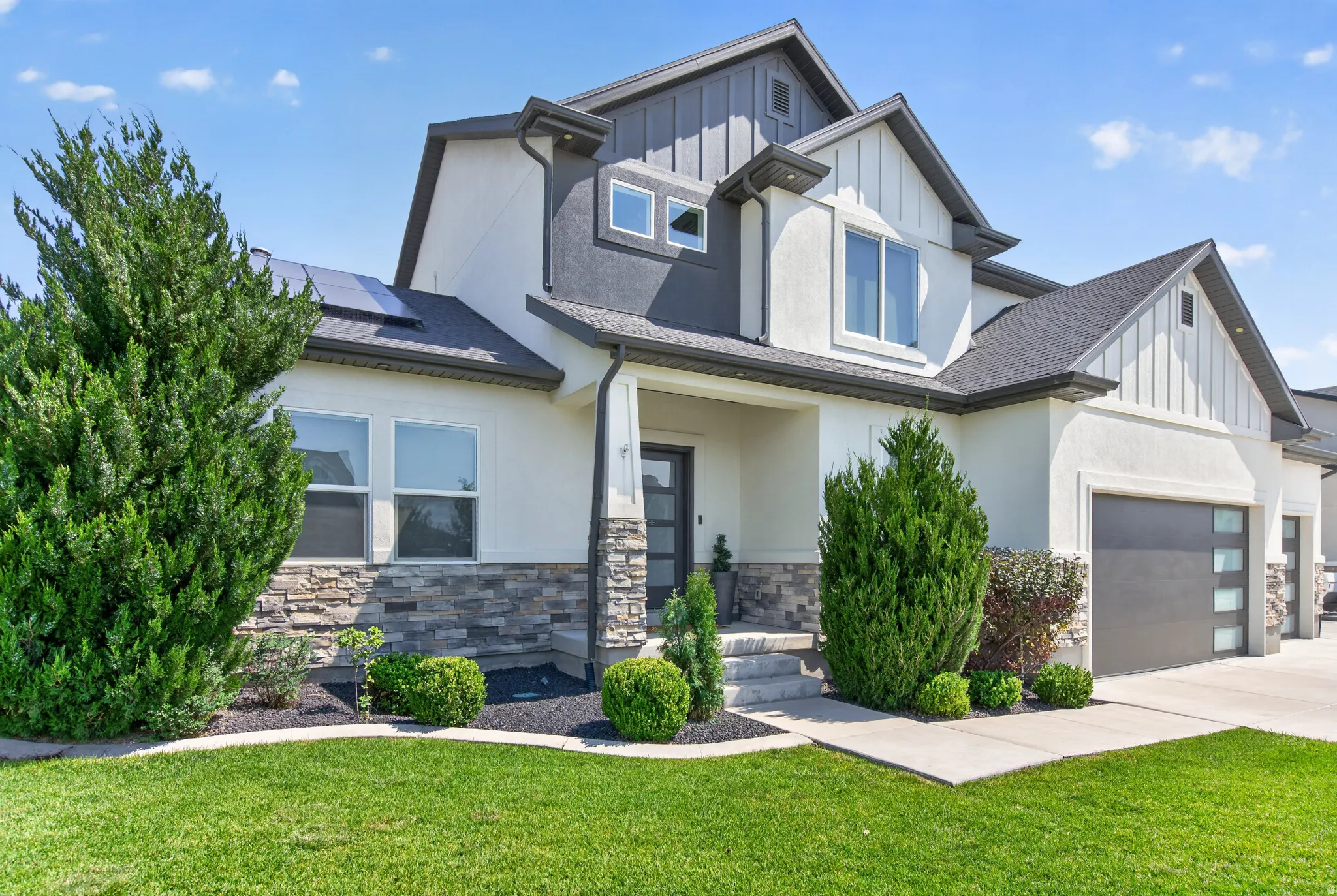 Modern stucco and stone facade featuring a dark gray garage door with frosted glass panels, an inviting entryway, and a verdant lawn with established landscaping