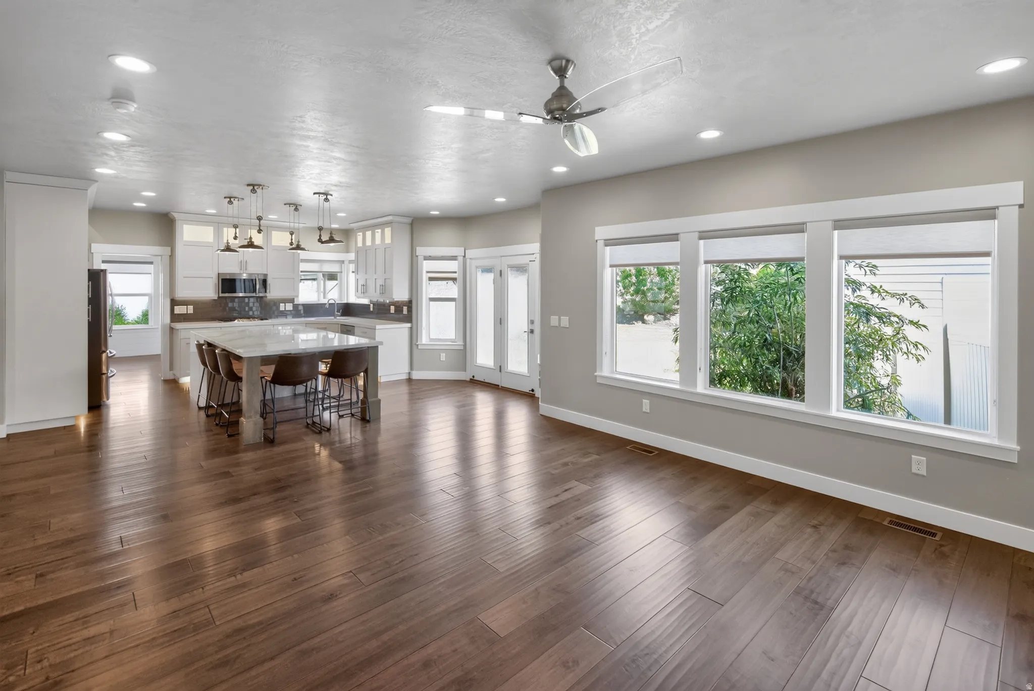 Open-concept living area featuring rich wood-finish flooring, a contemporary ceiling fan, recessed lighting, and a kitchen with white cabinetry, a large island, and pendant lighting