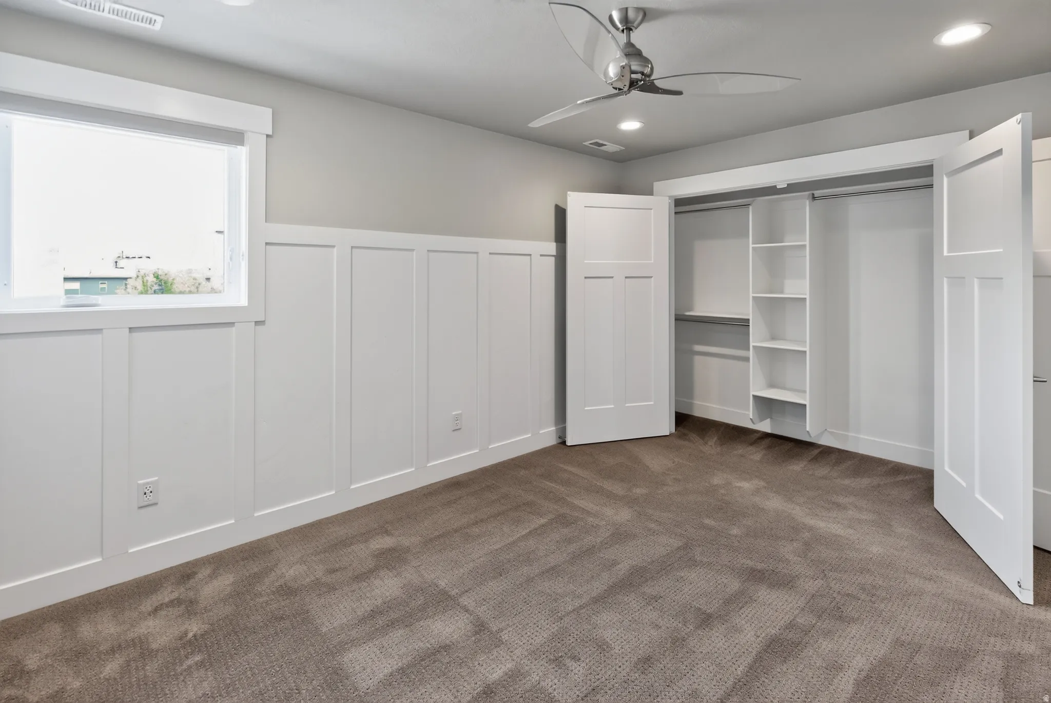 Carpeted room featuring a ceiling fan with integrated lighting, a window with white trim, board and batten wainscoting, and a closet with bifold doors and built-in shelving