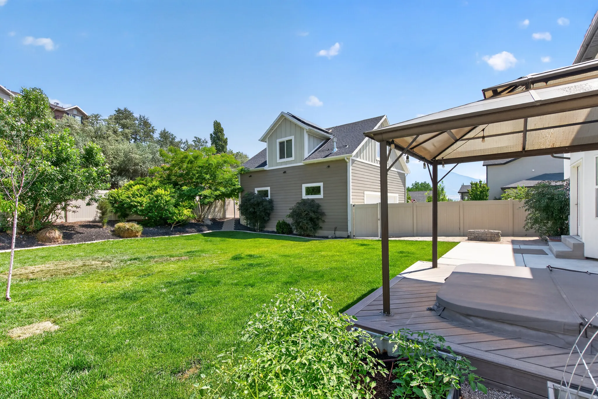 Expansive lawn area featuring a hot tub on a composite deck, a covered patio with a gazebo structure, and a concrete fire pit area