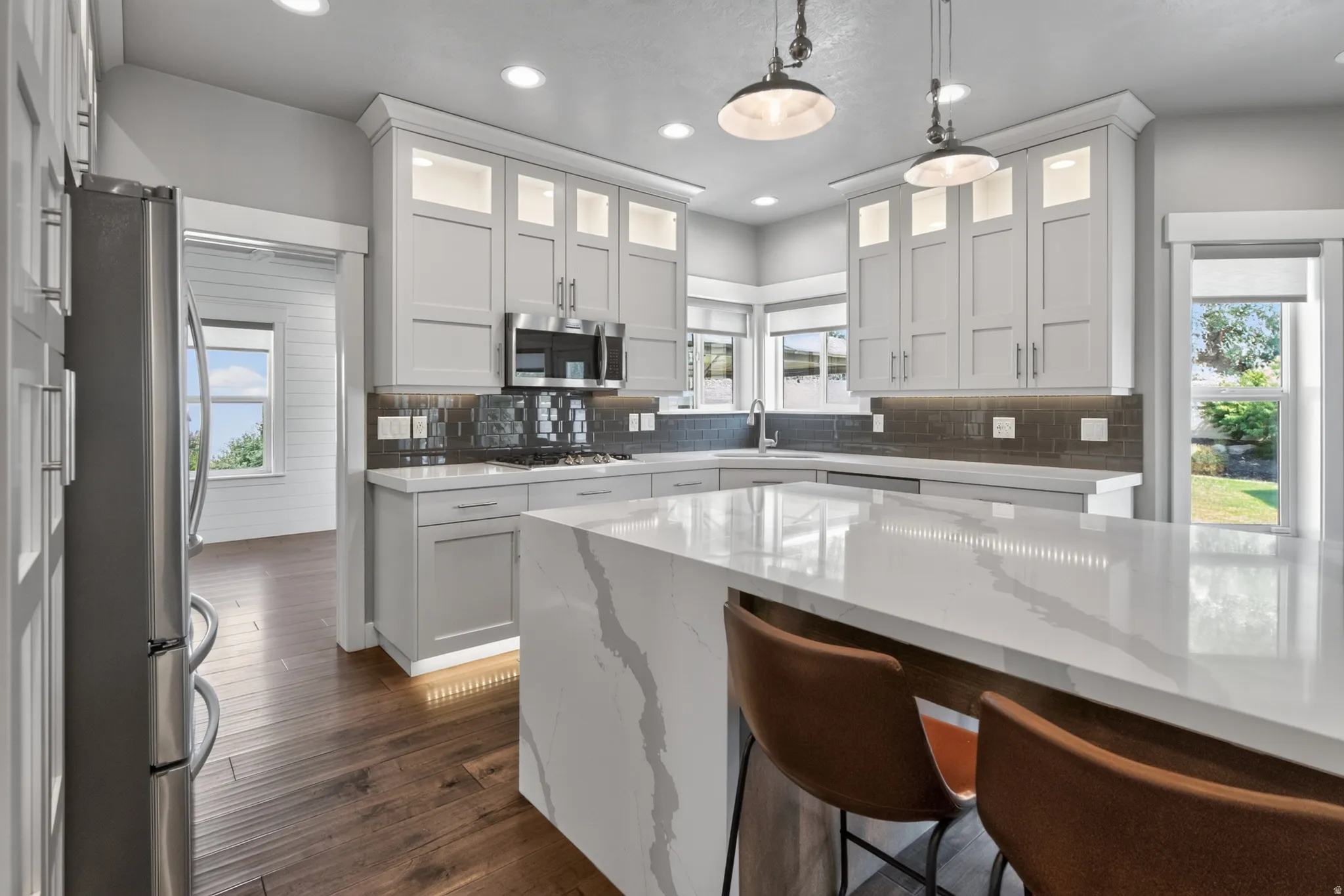 Kitchen featuring a waterfall-edge island with stone-finish countertop, light gray cabinetry with upper lighting, stainless steel appliances, and wood-finish flooring