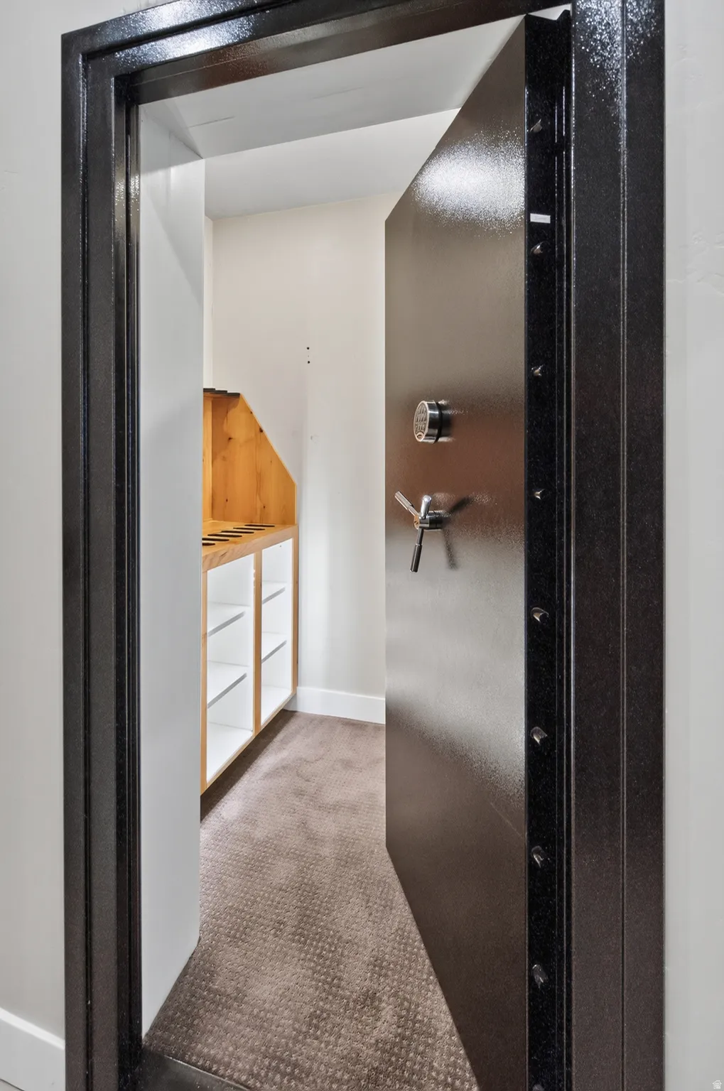 Heavy-duty vault door with a digital combination lock and handle, opening into a room featuring built-in wood shelving, white base trim, and patterned carpeting