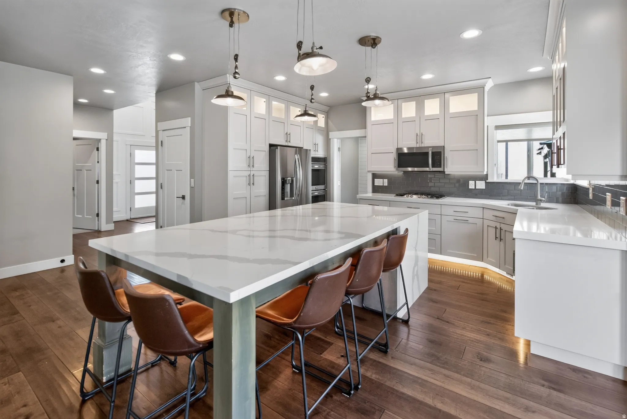 Spacious kitchen featuring a large island with a waterfall edge, white cabinetry with glass-front uppers, stainless steel appliances, a gray subway tile backsplash, and wood-finish flooring