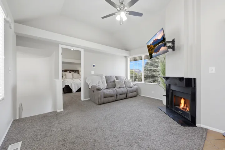 Living area featuring vaulted ceilings, a ceiling fan with integrated lighting, a built-in fireplace with a black surround, large windows with horizontal blinds, and a carpeted floor