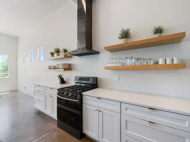 Modern kitchen featuring white cabinetry, light-toned countertops, a black range with an overhead range hood, and open wood-finish shelving