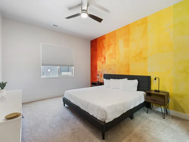 Interior room featuring a vibrant accent wall with a textured finish, light-colored carpeting, a ceiling fan with integrated lighting, and a window with a roller shade