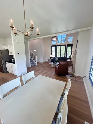 Wood-finish flooring throughout, kitchen with white cabinetry and dark countertops, modern chandelier, two-story living area with high windows, and a staircase with white balusters
