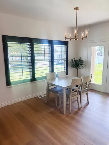Wood-finish flooring, dark-framed window with plantation shutters, bronze-finish chandelier with glass shades, white walls, and a white door with glass panels