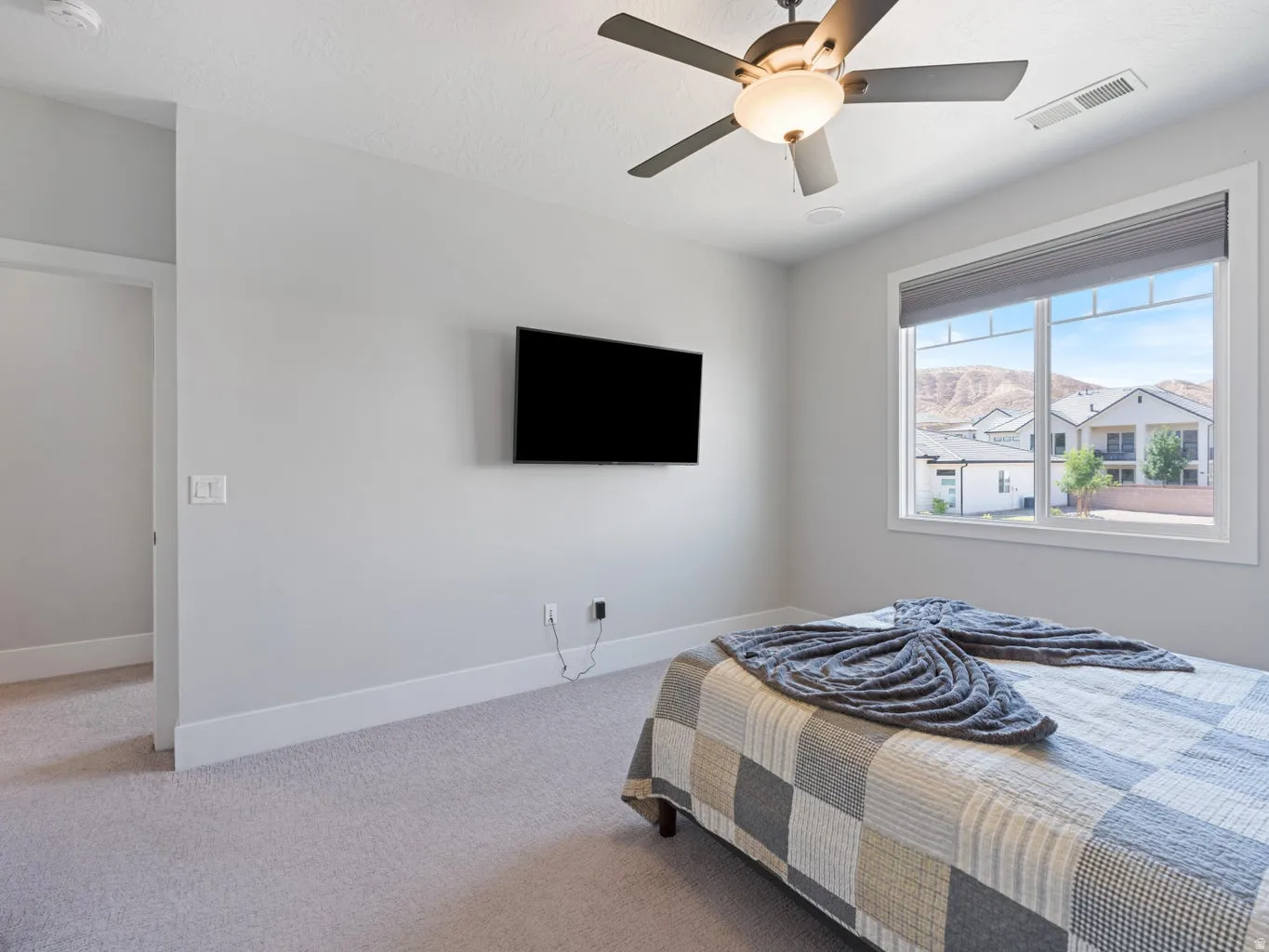 Bedroom featuring light gray walls, plush carpeting, a large window with roller shades, a ceiling fan with integrated lighting, and an installed flat-screen television