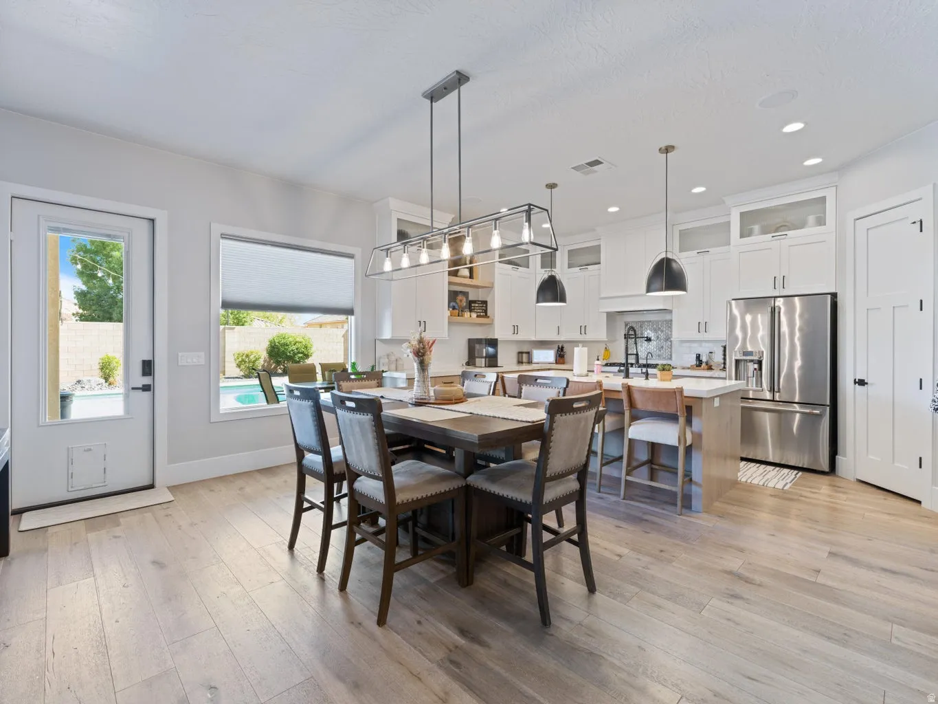 Open-concept kitchen and dining area featuring light wood-finish flooring, a contemporary linear chandelier, white shaker cabinetry with glass upper accents, stainless steel appliances, and a central island with counter seating