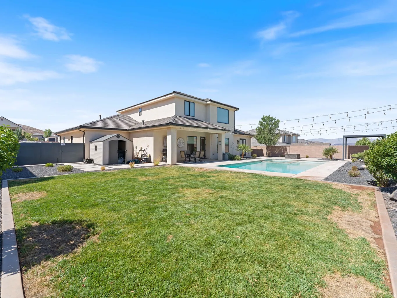 Expansive backyard featuring a rectangular swimming pool with surrounding concrete patio, a stucco exterior, an upper-level window, and a storage shed
