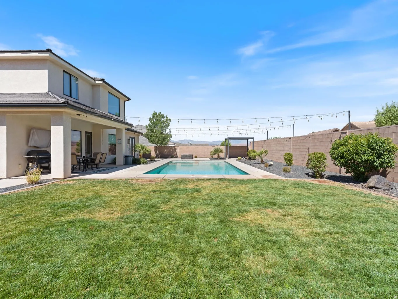 Expansive backyard featuring a manicured lawn, a rectangular swimming pool with surrounding hardscaping, a covered patio area, and perimeter string lighting