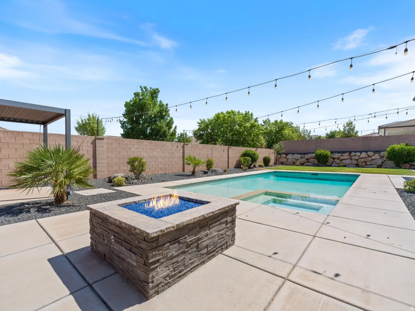 Backyard featuring a rectangular swimming pool with integrated steps, a stacked stone fire pit with blue glass media, an overhead string light installation, and a modern pergola