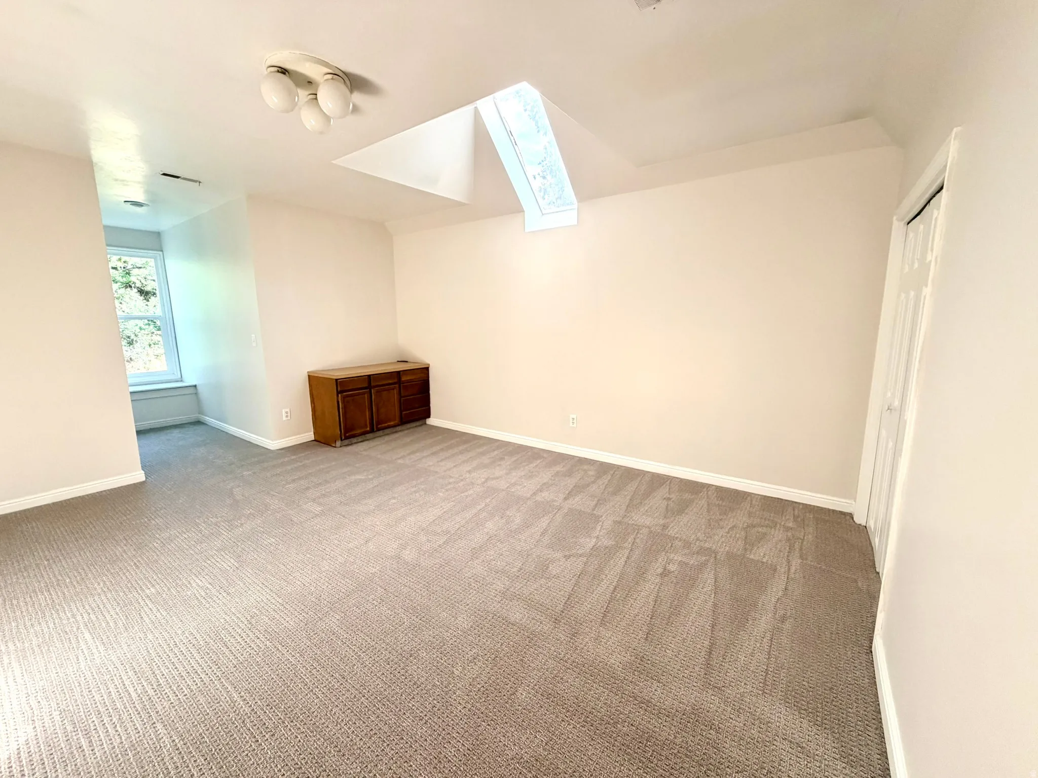 Spacious bedroom featuring neutral carpeting, a large window with natural light, a built-in wooden cabinet, and a ceiling-mounted light fixture