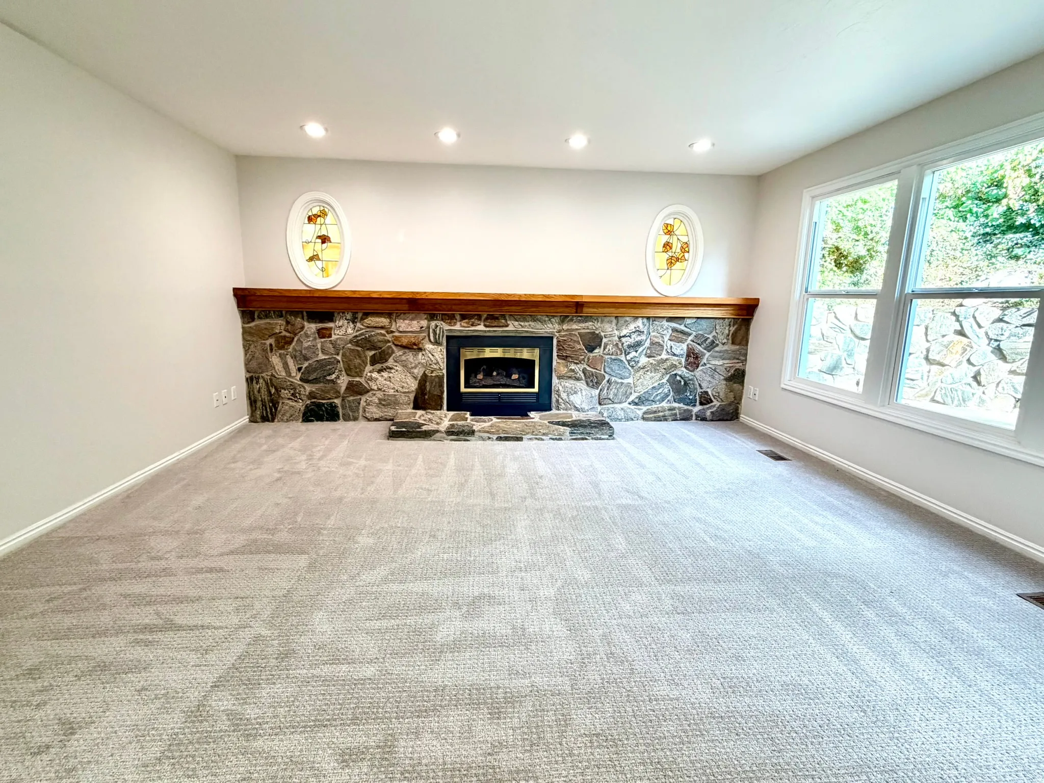 Spacious room featuring a stone-faced fireplace with a wood mantle, light-toned carpeting, recessed ceiling lighting, and large windows