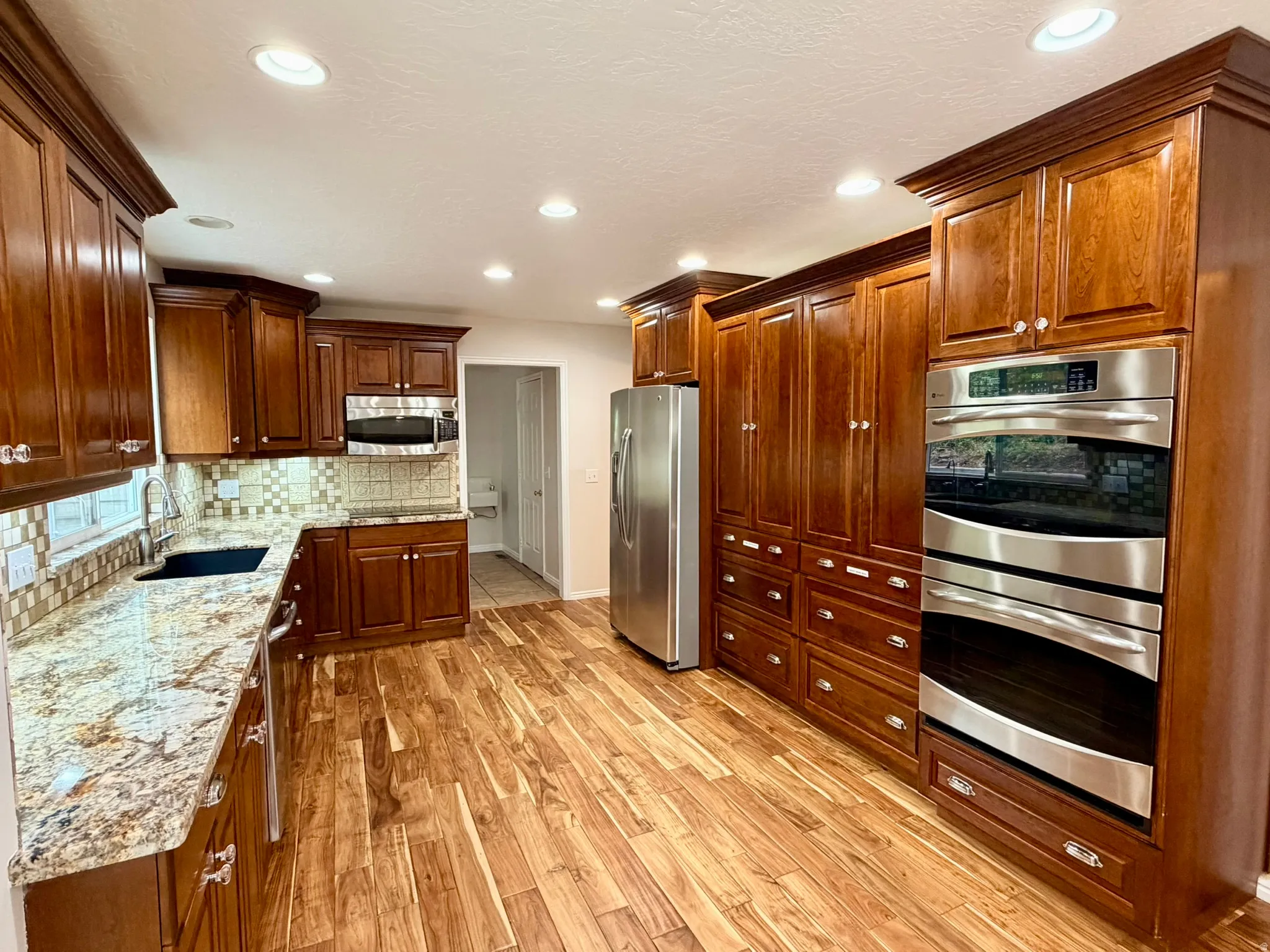 Gourmet kitchen featuring extensive wood cabinetry, granite countertops, stainless steel appliances including double ovens and a refrigerator, a tile backsplash, and wood-finish flooring