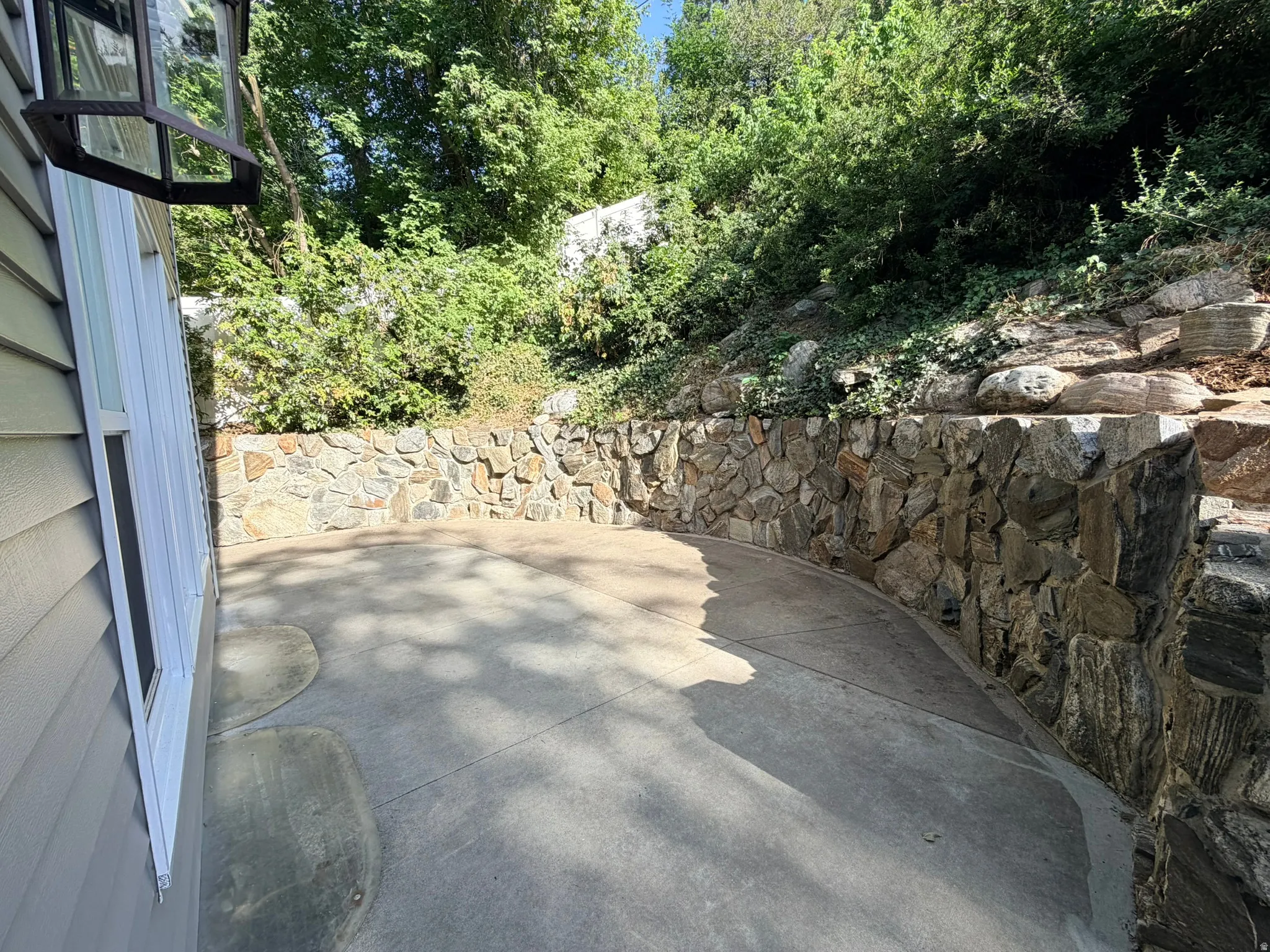 Spacious concrete patio featuring a curved stone retaining wall and surrounding mature foliage