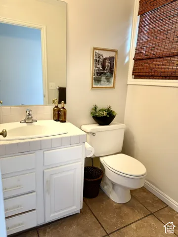 Vanity with white cabinetry and built-in sink, tiled countertop, large wall-mounted mirror, ceramic tile flooring, and a toilet