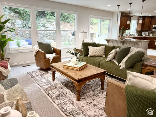 Expansive windows with white trim, stone fireplace hearth, wood-finish coffee table, recessed lighting, and kitchen island with pendant lighting