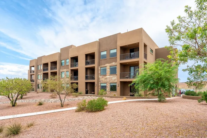 Contemporary multi-story building featuring stucco exterior, stone accents, multiple balconies with dark railings, and a gravel landscape with desert plantings