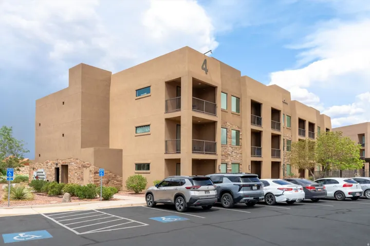 Multi-story stucco building featuring stone veneer accents, individual unit balconies, prominent building numbering, and a dedicated parking area with marked accessible spaces