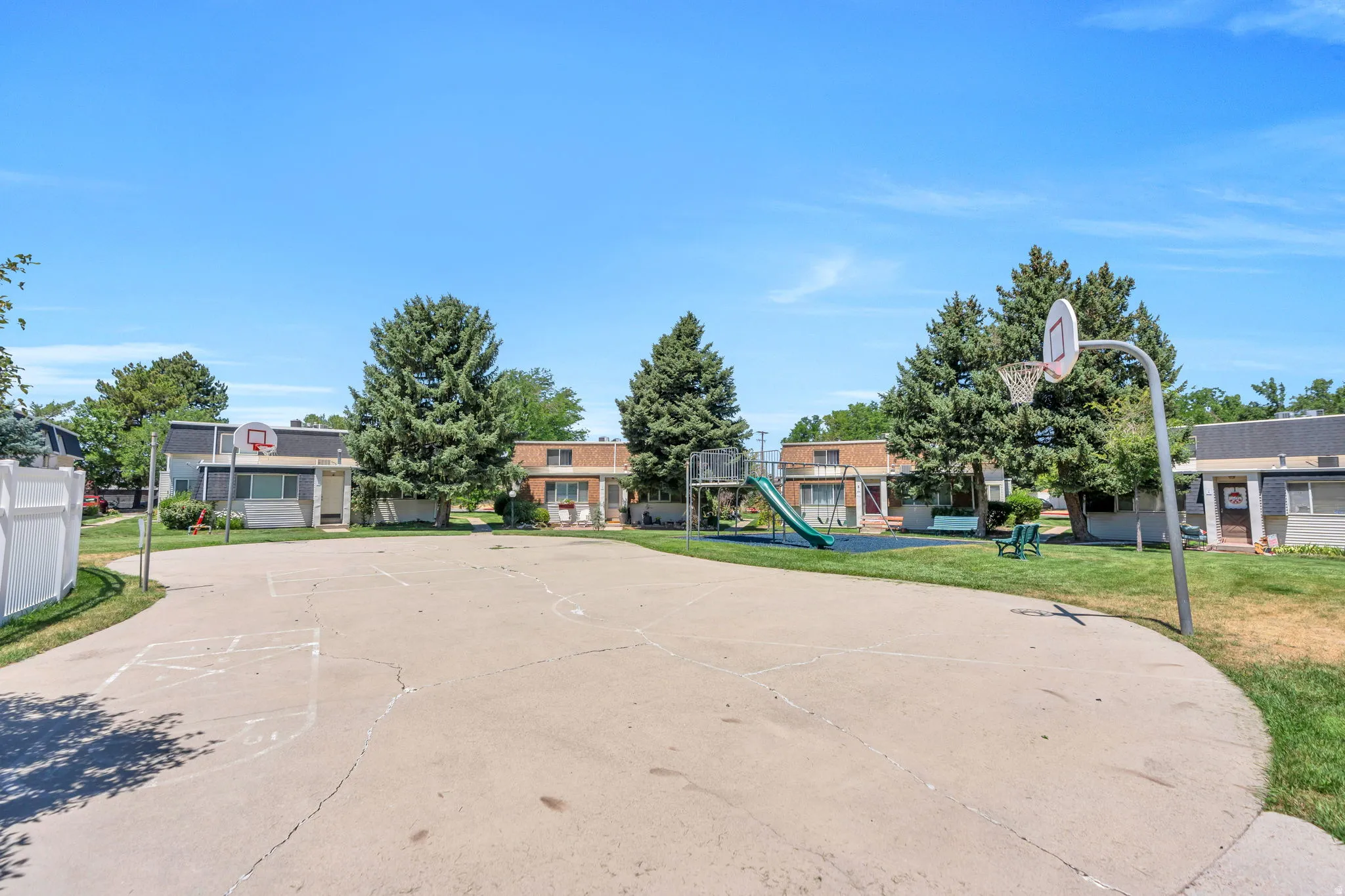 Paved outdoor court featuring a basketball hoop, white privacy fencing, and mature trees