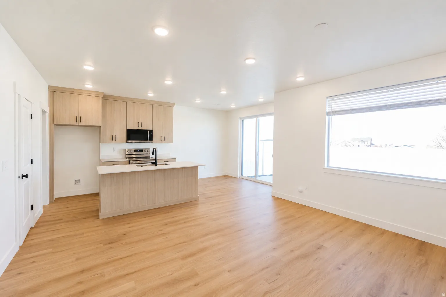 Open-concept living area featuring wood-finish flooring, a kitchen island with a sink, light wood-finish cabinetry, and stainless-steel appliances