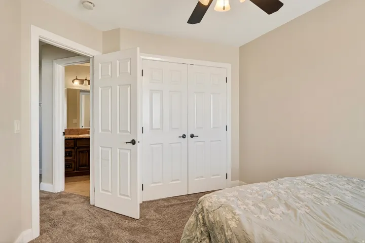 Bedroom space featuring neutral wall paint, textured carpet flooring, a ceiling fan with integrated lighting, and white paneled doors leading to a closet and a bathroom with a dark wood vanity and light-toned countertop