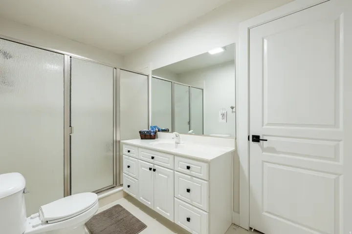Bathroom featuring a single vanity with stone-finish countertop, white cabinetry with black hardware, a shower with frosted glass doors, a toilet, and a paneled interior door