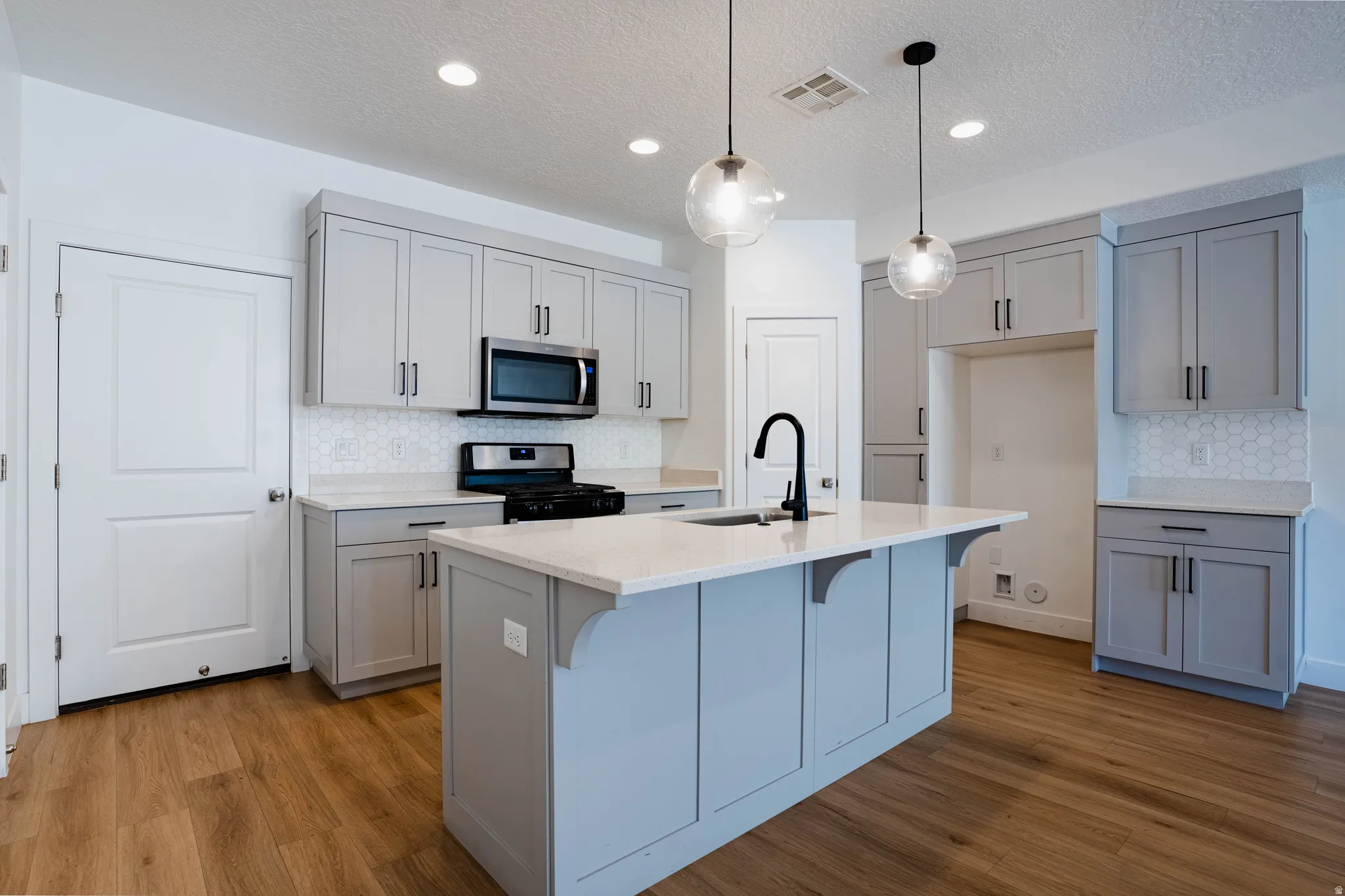 Kitchen featuring a central island with an integrated sink and matte black faucet, light gray cabinetry, stainless steel microwave, gas range, and wood-finish flooring