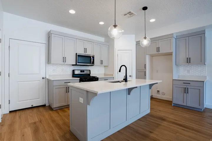 Kitchen featuring a central island with an integrated sink and matte black faucet, light gray cabinetry, stainless steel microwave, gas range, and wood-finish flooring