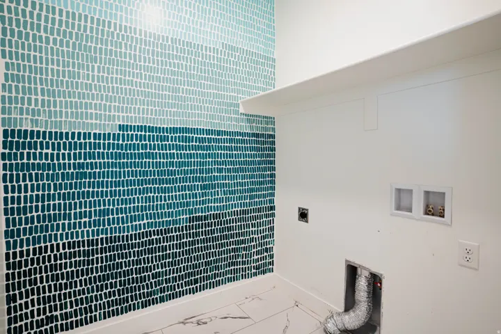Laundry area featuring a multi-toned blue mosaic tile accent wall, white wall shelving, white electrical outlets, and a floor with a light-colored marble-look pattern