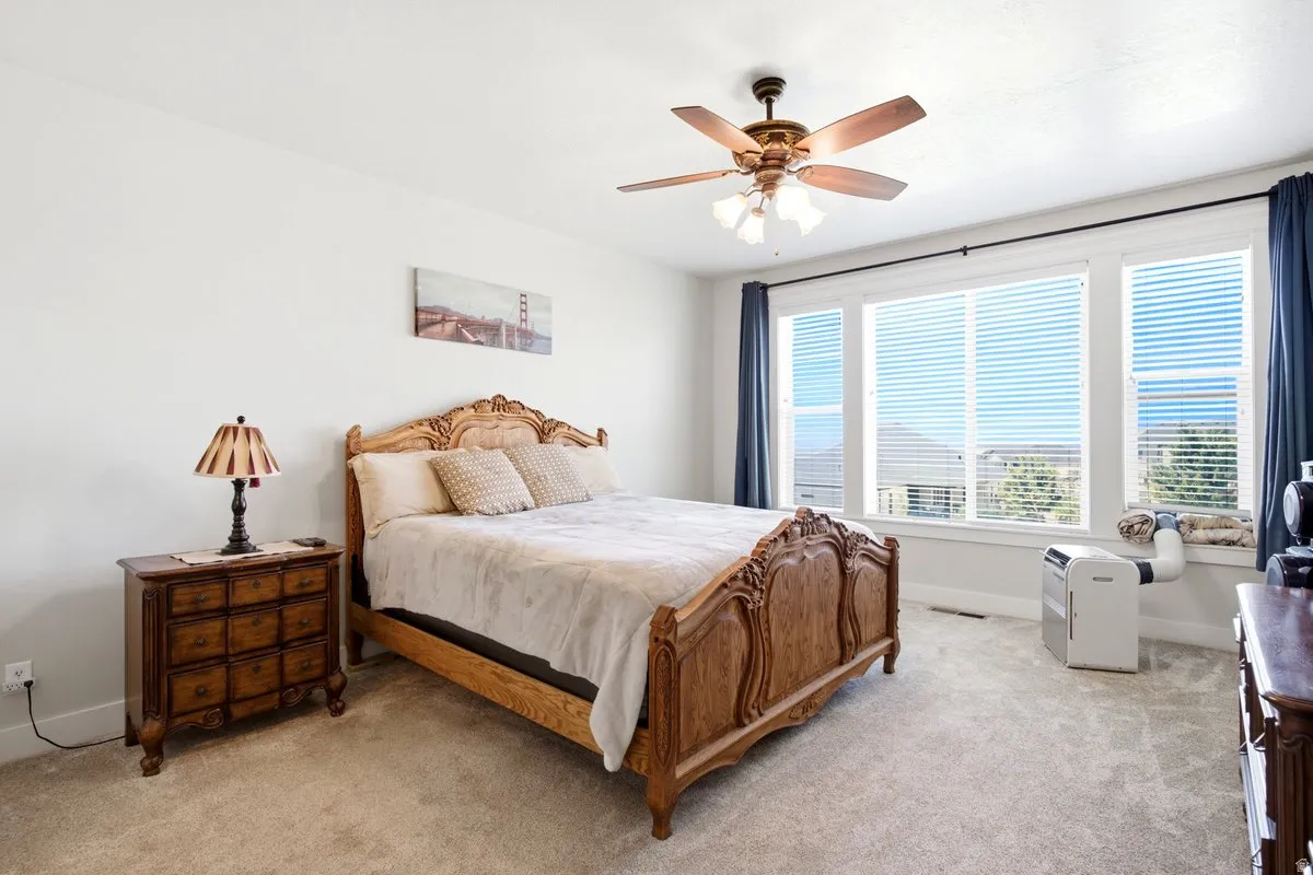 Spacious room featuring carpeting, multiple windows with blinds and drapes, a ceiling fan with integrated lighting, and white walls
