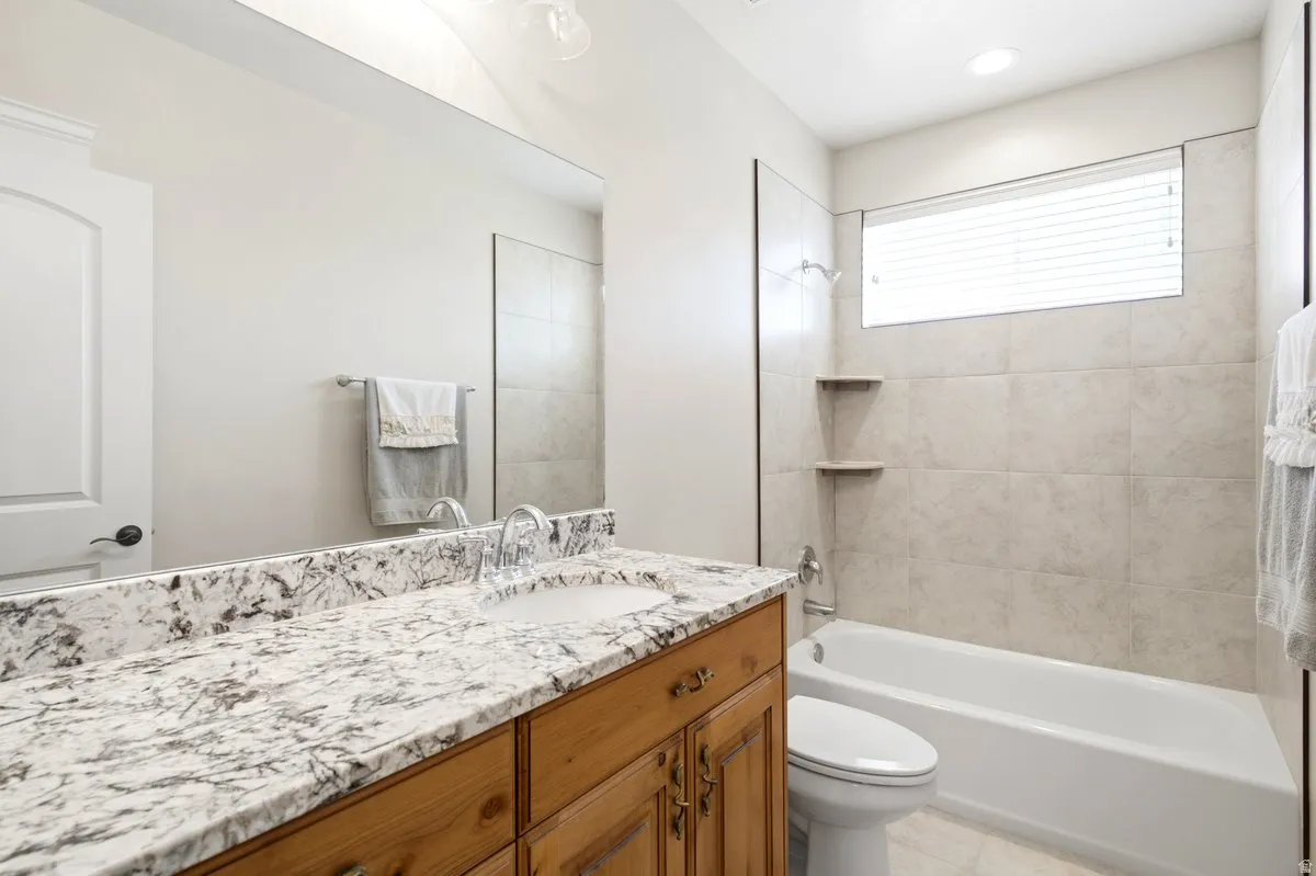Bathroom featuring a wood-finish vanity with a built-in sink and stone countertop, a full-width wall mirror, a bathtub with tile surround, and a high-set window with blinds