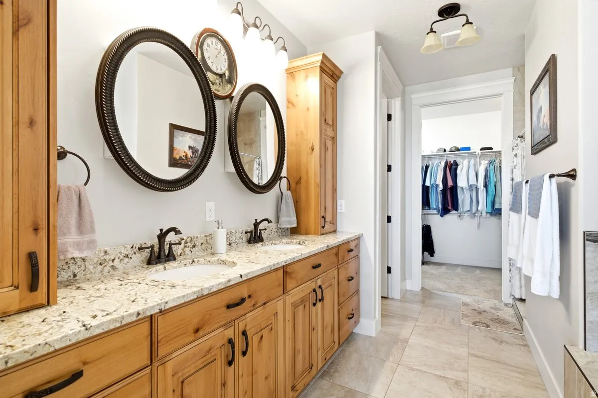 Double vanity with light-colored speckled countertops, twin oval mirrors with dark frames, wood-finish cabinetry, and a walk-in closet beyond